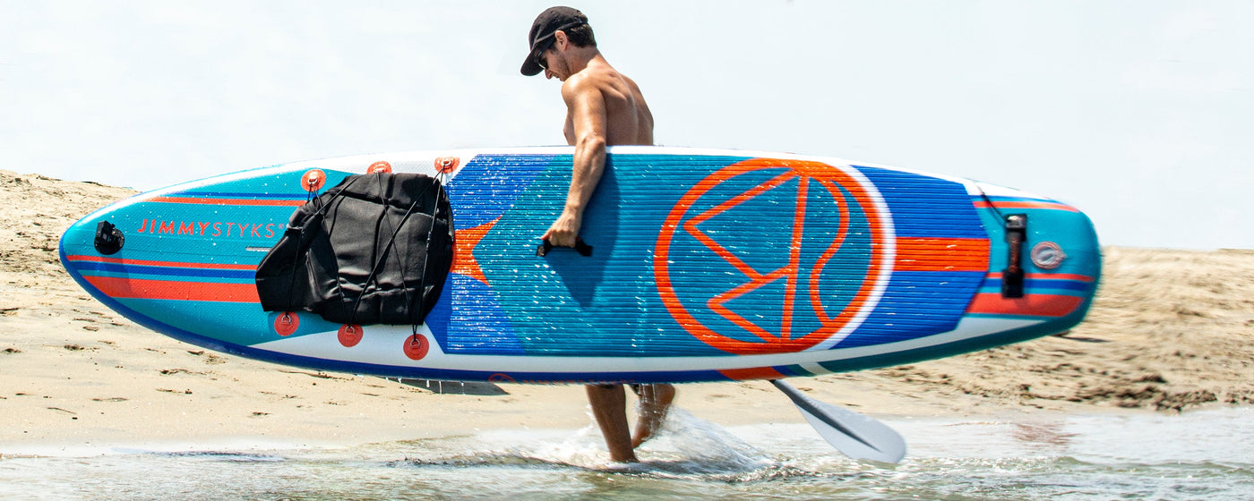A man is holding Jimmy Styks Inflatable SUP in one arm on the beach